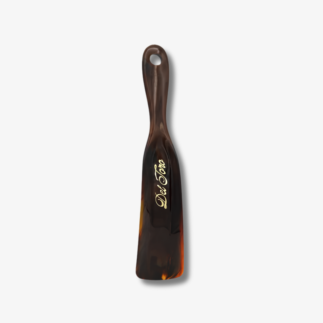 The Del Toro Shoes “Del Toro Shoe Horn - Tortoiseshell” is a brown tortoiseshell-patterned shoe horn, featuring a convenient hole in the handle and “Del Toro” elegantly written in gold cursive. This travel-sized accessory is shown against a plain white background.