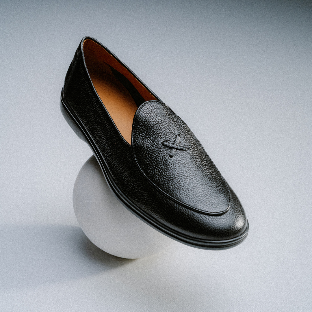 The Men’s Black Pebbled Leather Viaggio Loafer by Del Toro Shoes, featuring refined pebbled Italian leather and subtle stitching, is elegantly poised atop a sleek white sphere against a gentle gradient background, capturing the spirit of stylish comfort in a casual shoe.