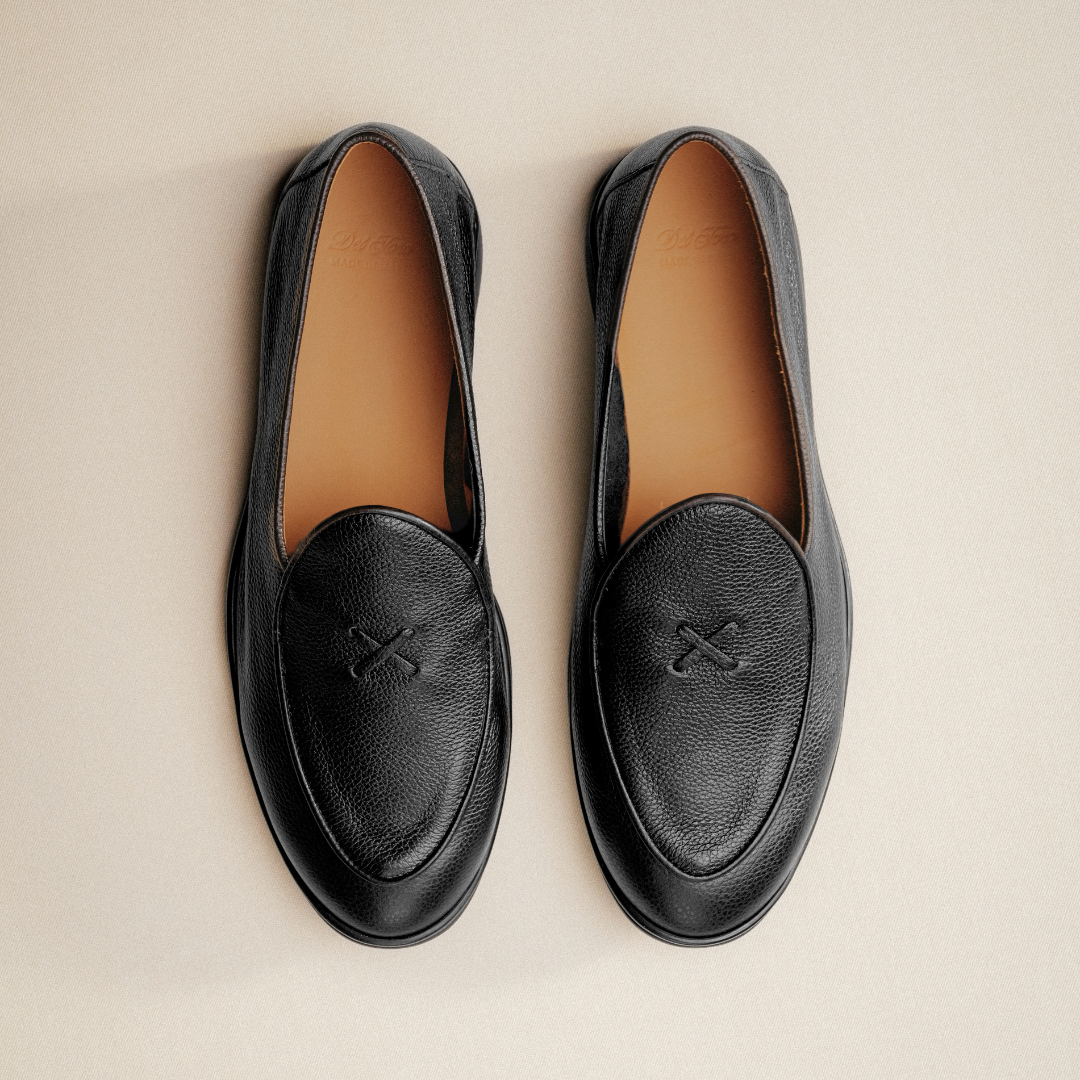 A pair of Del Toro Shoes Men’s Black Pebbled Leather Viaggio Loafers with tan insoles, viewed from above, showcasing slightly rounded toes and distinctive crossed stitching on the top of each shoe, set against a light beige background—an ideal choice for comfortable casual wear on any occasion.