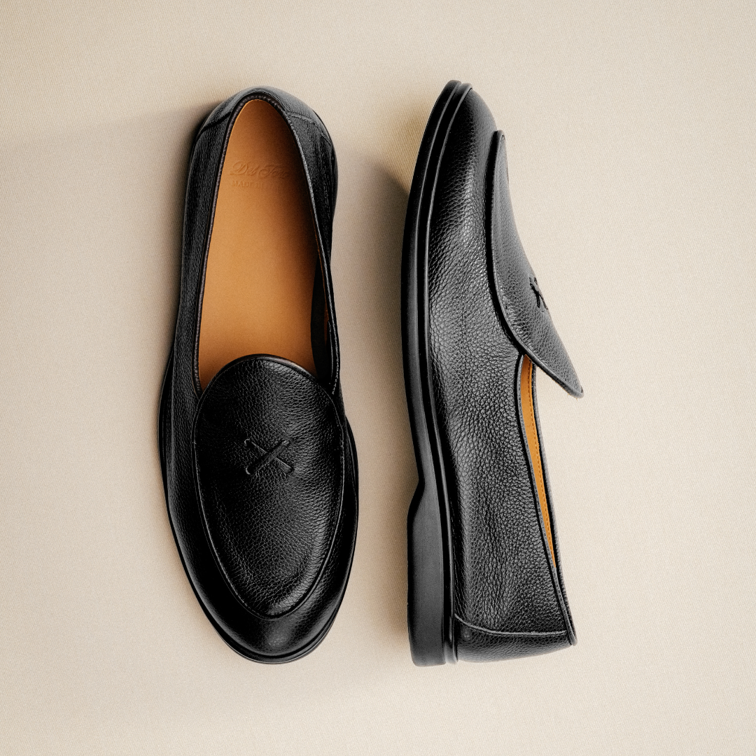 A pair of Men’s Black Pebbled Leather Viaggio Loafers from Del Toro Shoes, featuring tan insoles, is displayed on a light beige background. One loafer stands upright while the other rests on its side to showcase its sole and profile—an elegant and versatile casual shoe designed for timeless comfort on any occasion.