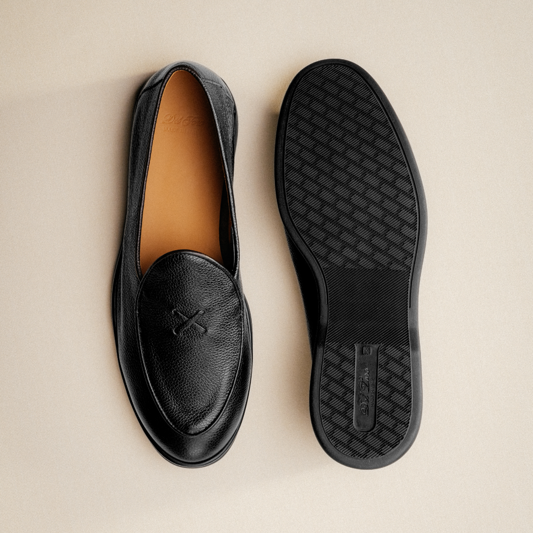 A pair of Men’s Black Pebbled Leather Viaggio Loafers by Del Toro Shoes is displayed on a beige surface—one shoe upright showcasing the stitched detail on the top, and the other flipped to reveal its textured sole—offering a sophisticated yet comfortable casual option.