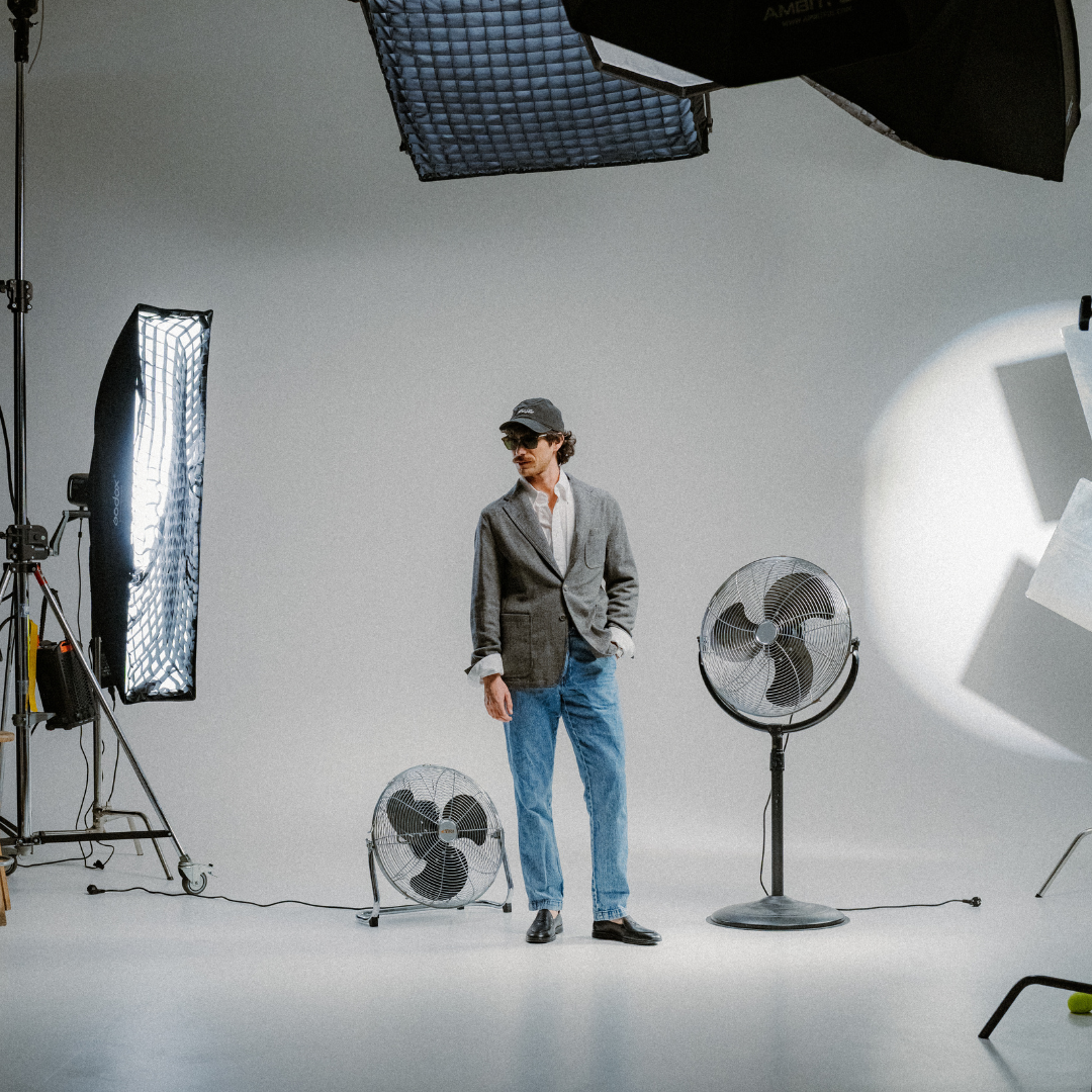 A man in a blazer, jeans, and cap stands in a minimalist photography studio with large lights and two fans—one on the table and one on the floor—surrounding him. Completing his look is the Del Toro Shoes Men’s Black Pebbled Leather Viaggio Loafer, a comfortable casual shoe crafted from Italian leather. The setting features a clean white background.