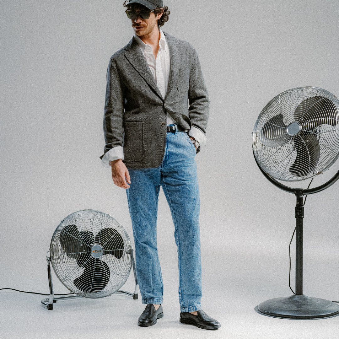 A man in a gray blazer, white shirt, blue jeans, and Del Toro Shoes’ Men’s Black Pebbled Leather Viaggio Loafers stands between two electric fans against a plain background. His comfortable Del Toro loafers and sunglasses complete the effortlessly cool look.