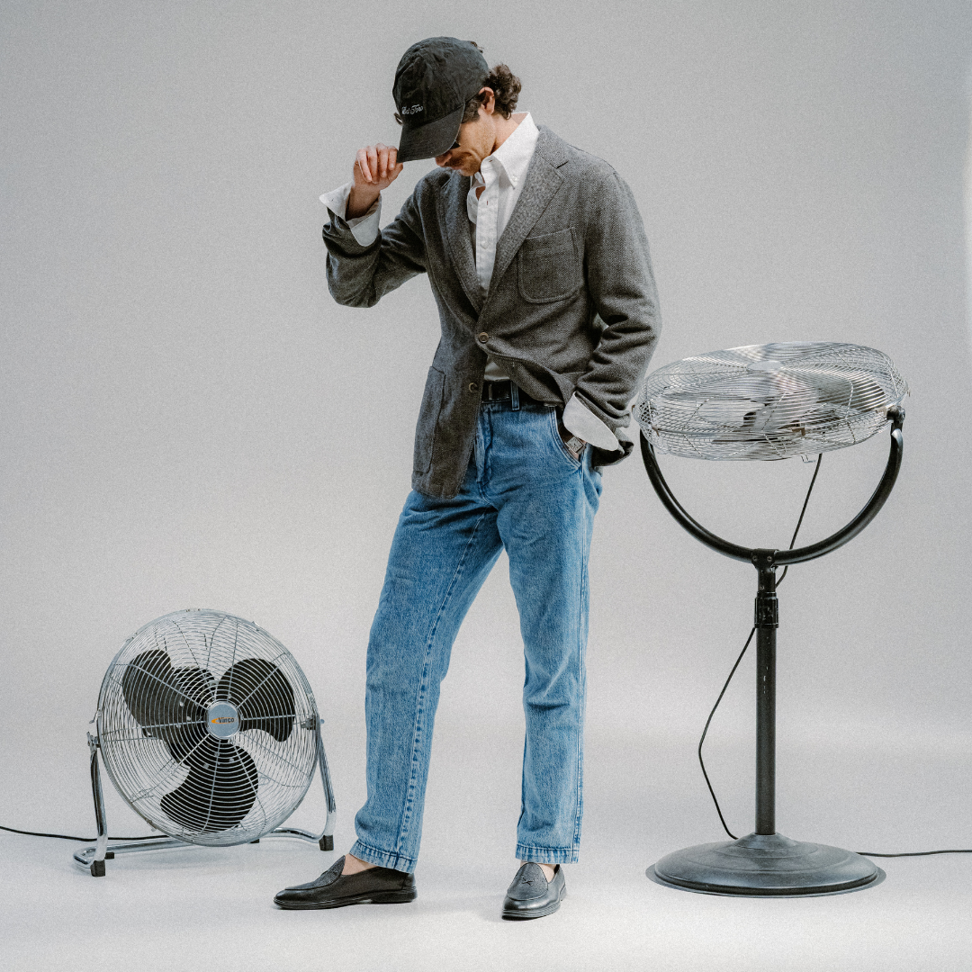 A person in a blazer, jeans, and cap stands between two metal fans in a studio with a plain gray background, effortlessly completing their look with the Men’s Black Pebbled Leather Viaggio Loafer from Del Toro Shoes—a comfortable casual shoe that adds understated elegance to the ensemble.