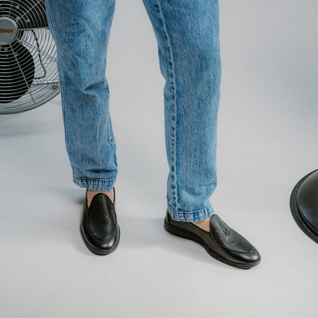 A person wearing blue jeans and the Del Toro Shoes Men’s Black Pebbled Leather Viaggio Loafer, a comfortable casual shoe crafted from fine Italian leather, stands on a light-colored floor. Part of a metal fan and the base of a lamp are visible on either side of their legs.